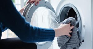 Homeowner redistributing clothes inside a front-loading washing machine while inspecting the drain hose and filter in a clean laundry room.